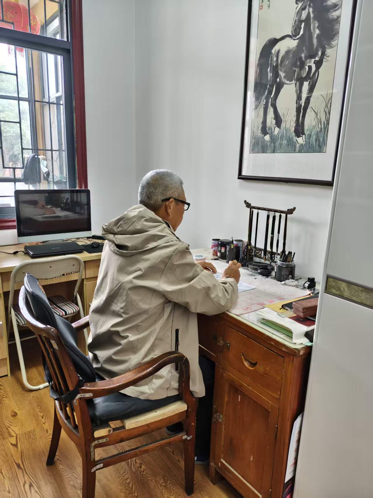 Qing Wu seated at a wooden desk, with daylight from a window and a painting above the brushes